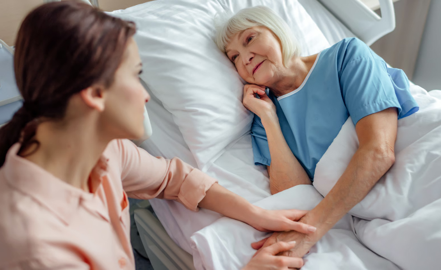 A young woman in a peach shirt sits by a hospital bed, gently holding the hand of an elderly woman with white hair who is looking at her peacefully.