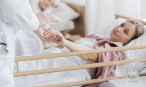 A physician in a white coat gently holds the hand of a blonde woman lying in a hospital bed, who is looking at him with a focused, calm expression.
