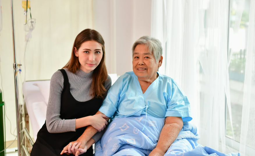 A young woman sits supportively on a hospital bed, holding the hand of an elderly woman in a blue gown, illustrating the emotional connection and grief support provided during hospice care.