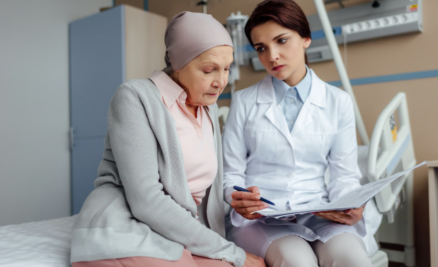 A female doctor points to details in medical documents while an elderly patient wearing a light pink headscarf listens intently on a hospital bed.