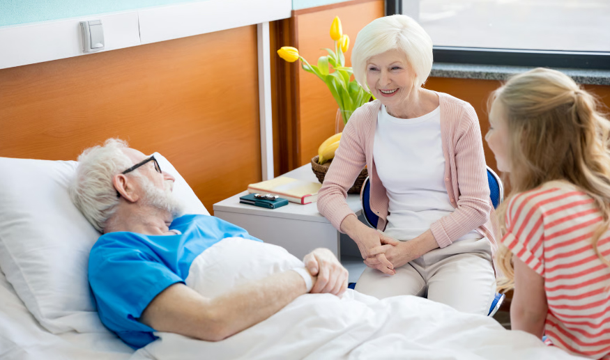 An elderly man in a hospital bed smiling and talking with his wife and granddaughter during a hospice facility visit.