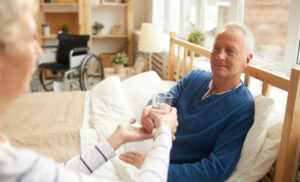 An elderly man sitting up in bed receiving a glass of water from a caregiver, illustrating comfort care in a home setting.