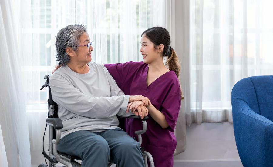A young Asian nurse in purple scrubs kneels down to comfort and talk with an elderly senior woman sitting in a wheelchair in front of a window.