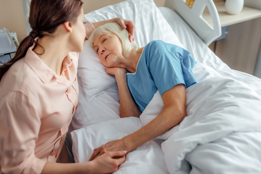 A young woman lovingly strokes the head and holds the hand of an elderly woman sleeping peacefully in a hospital bed.