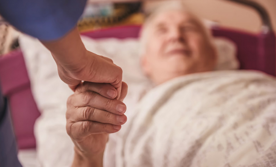 A close-up of a hospice caregiver gently holding the hand of an elderly patient resting in a hospital bed, shifting the focus from food to emotional connection.