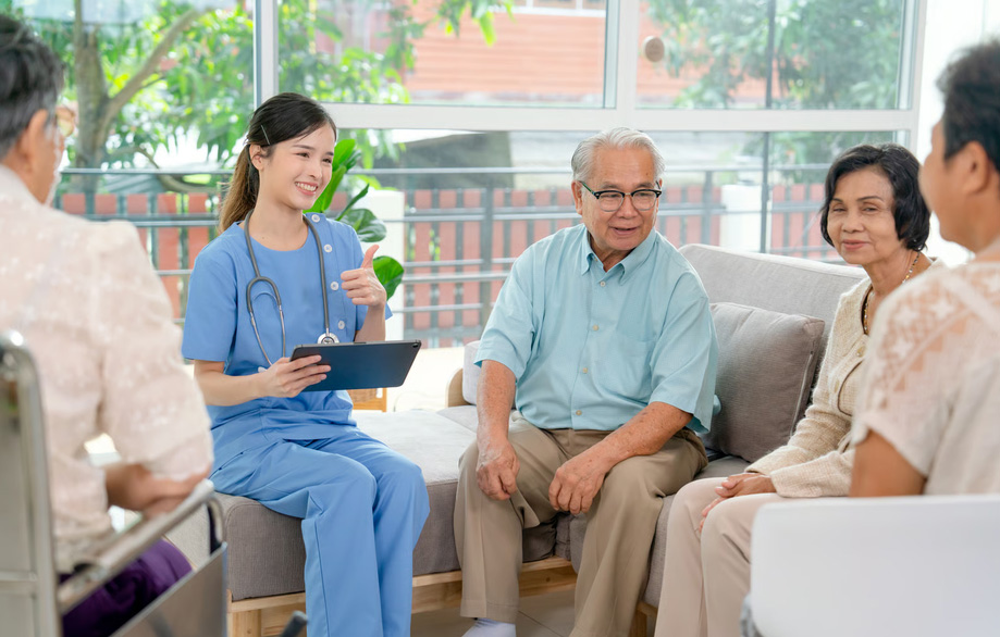 A smiling young female hospice nurse in blue medical scrubs holding a tablet and giving a thumbs-up during a reassuring conversation with an elderly man and his family members on a living room sofa, illustrating the compassionate professional support provided during end-of-life care.