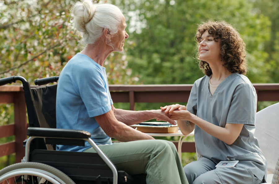 An elderly woman with white hair in a hair bun sits in a wheelchair on a wooden deck, smiling and holding hands with a female caregiver in grey scrubs against a background of green trees.