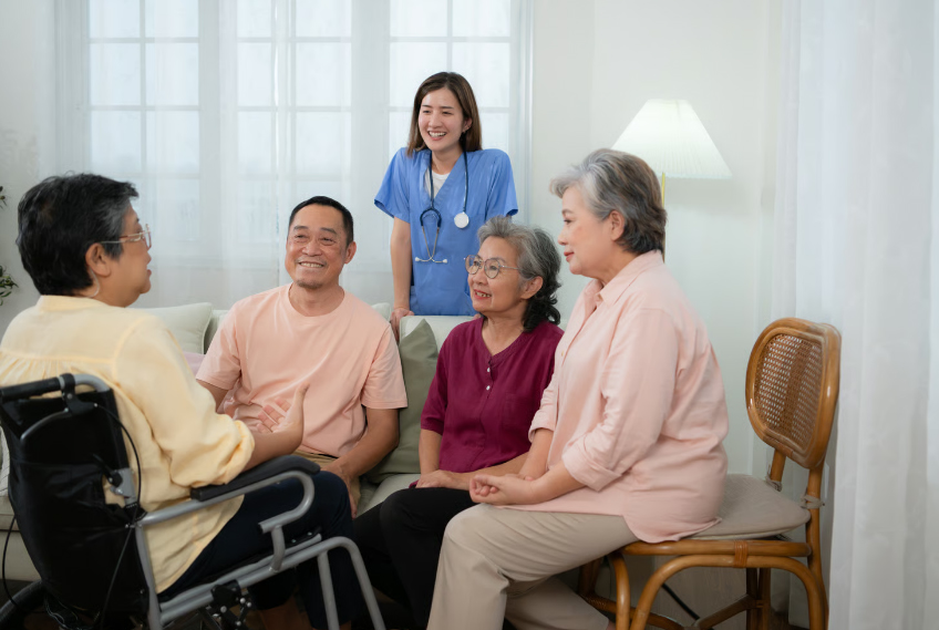 A group of four smiling Asian seniors sitting in a bright living room, including a woman in a wheelchair, talking together with a nurse in blue scrubs standing behind them.