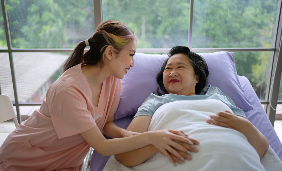 A young female home caregiver in pink scrubs holding hands and smiling at an elderly Asian woman resting in a hospital bed.