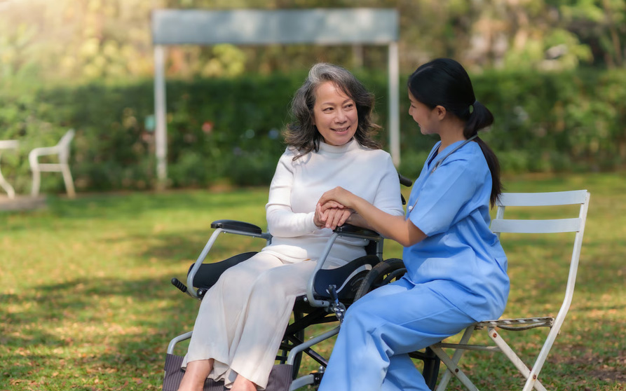 A hospice nurse in blue scrubs sits next to a senior woman in a wheelchair, smiling and holding her hand in a sunny outdoor park setting.