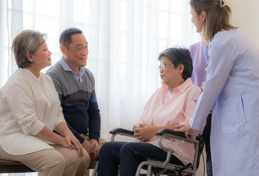 A healthcare professional talking with an elderly woman in a wheelchair and her family members in a bright room.