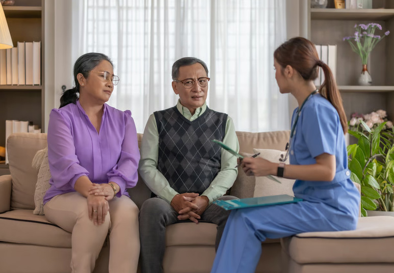 A hospice admission nurse in blue scrubs holding clipboard documents while consulting with a senior couple on their living room sofa during the first 72 hours of hospice care.