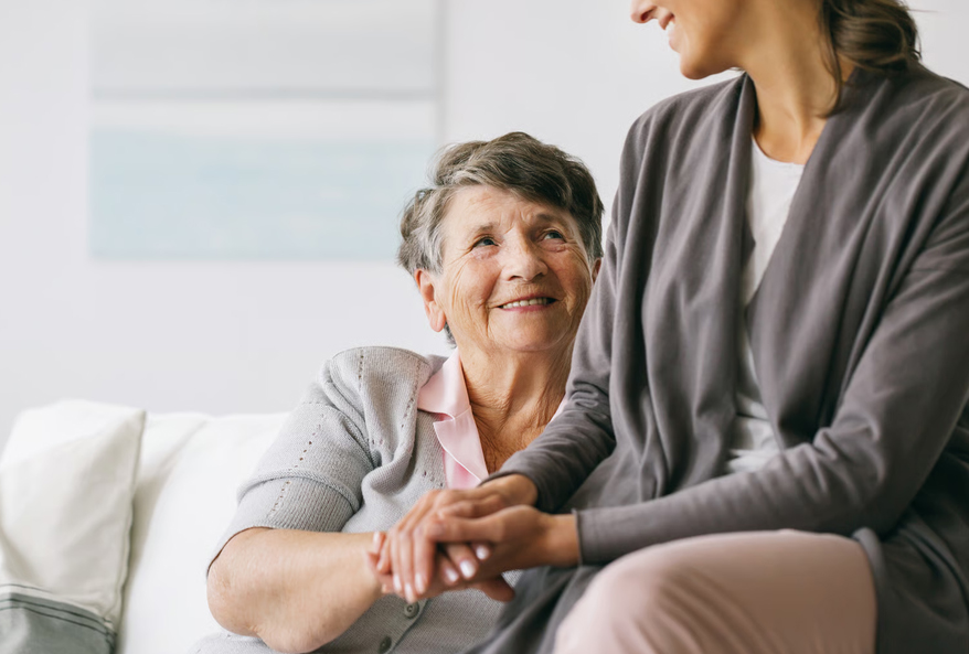 An elderly mother sitting on a couch and smiling while holding hands with her adult daughter.