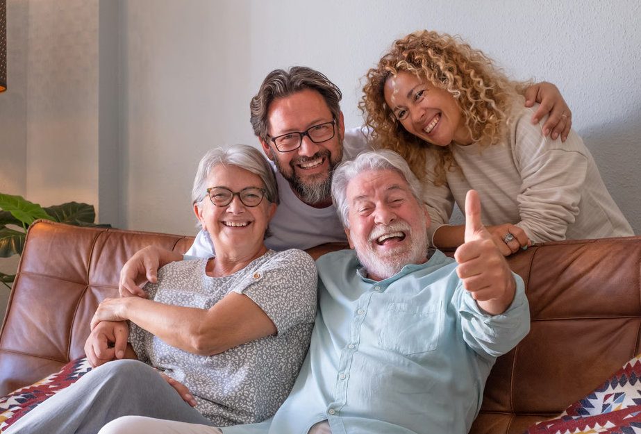A happy multi-generational family smiling together on a sofa, with a senior man giving a thumbs-up during a hospice family support gathering in Atlanta.