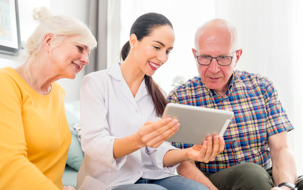 A female hospice healthcare professional reviewing medical information on a digital tablet with a senior patient and his family member during a recertification visit.