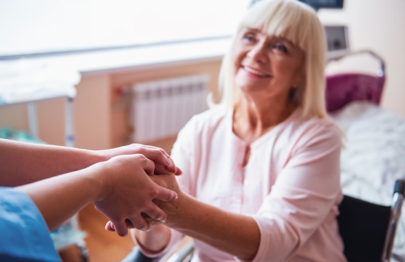 A caring hospice caregiver in blue scrubs holding the hands of a smiling elderly woman, illustrating the compassionate, Medicare-covered support provided to families in Metro Atlanta.