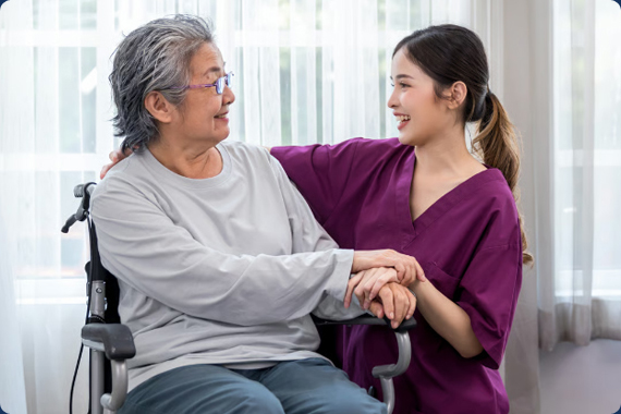 A young female caregiver in purple scrubs smiling and holding hands with an elderly Asian woman in a wheelchair inside a bright senior care facility